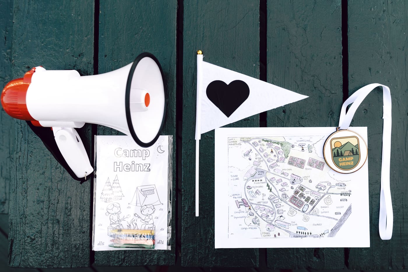 Camp Heinz summer camp themed wedding weekend flat-lay: megaphone, pennant flag with heart, hand-drawn camp map, custom Camp Heinz coloring book and lanyard medallion